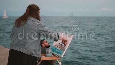 在海边的一对年轻漂亮的夫妇。 有创意的女人画这幅画，男人弹吉他。